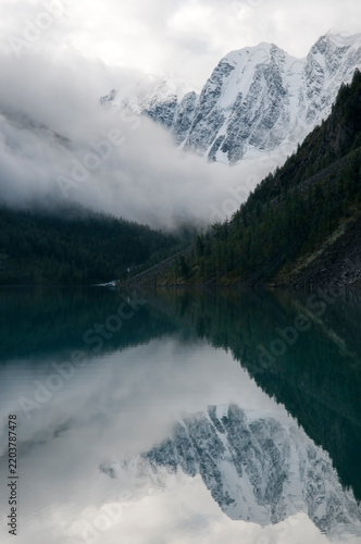 Early morning view of the Lower Shavlinskoye lake with mountain peaks Dream, Fairy Tale and Beauty covered in clouds and mirror themselves in the water, Russian Federation