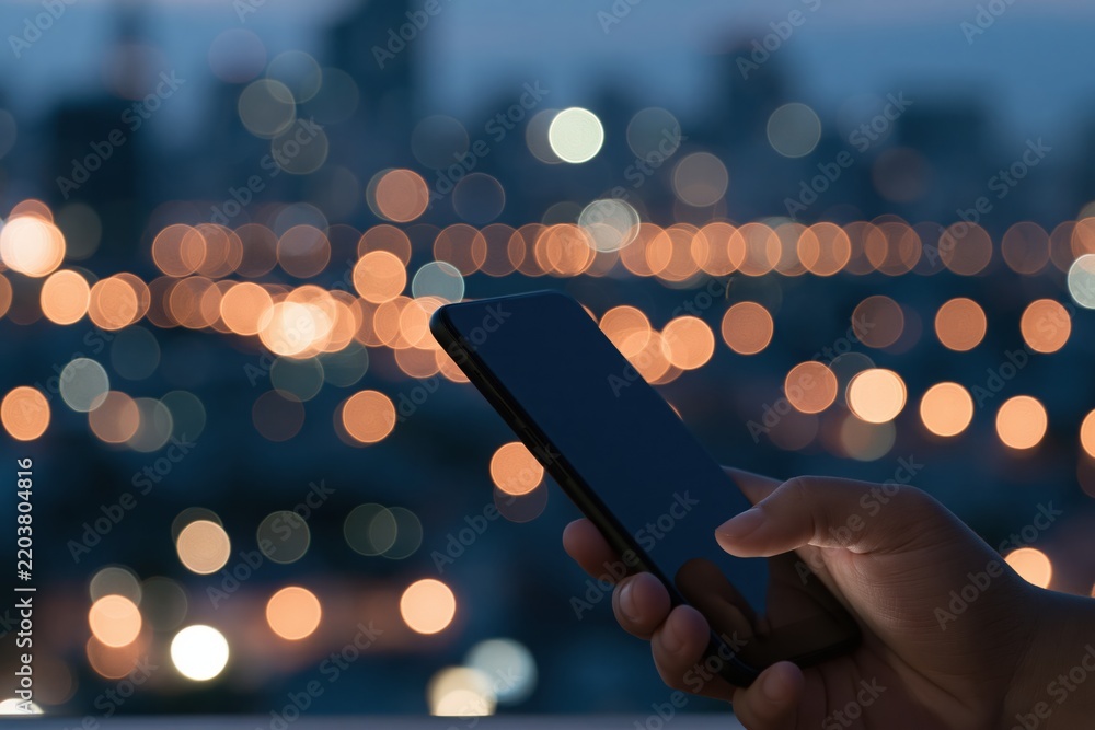Fototapeta premium Hand holding smartphone against city night lights bokeh