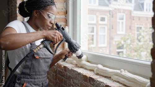 Construction worker applying expanding foam around window frame before smoothing insulation along a brick wall, protective eyewear and gloves, interior renovation footage