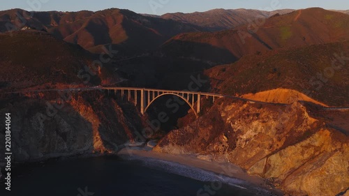 Aerial view captures Bixby Creek Bridge along the rugged Big Sur coast in California during golden hour sunset, showing coastal cliffs, beach sand, and ocean surf.