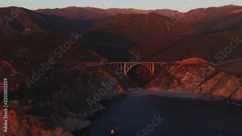 Aerial view of Bixby Creek Bridge spanning the rugged canyon along the Pacific coast in Big Sur, California during warm sunset illumination. Ocean waves lap the sandy cove below the arch structure.