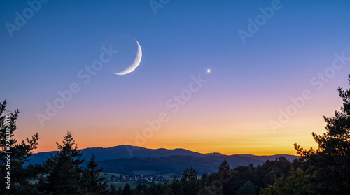 Fina luna creciente acompañada de una estrella vespertina en un cielo despejado durante el crepúsculo, con suave degradado de azul a tonos cálidos dorados.