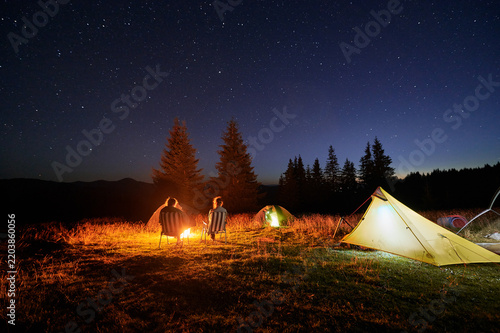Two people campers sit by campfire under starry sky, surrounded by illuminated tents. Serene, clear night in mountains creates peaceful atmosphere, perfect for stargazing and relaxing in nature.
