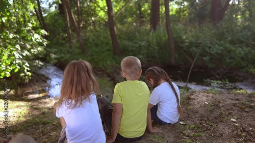 children sit on the banks of river. happy family lifestyle childhood dream concept. a group of little children sit together by the river and talk. kids spend time outdoors in nature in the forest