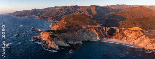 Aerial wide perspective of Bixby Creek Bridge spanning over a steep coastal canyon along the Pacific coastline in Big Sur, California during warm sunset illumination.