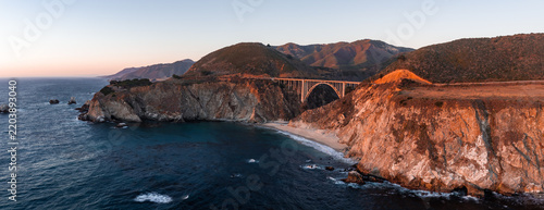 Aerial view showcases Bixby Creek Bridge spanning the rugged coastline of Big Sur in California during sunset, where cliffs drop down to the Pacific Ocean shoreline.