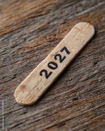 A popsicle stick reading 2027 resting on a wooden surface