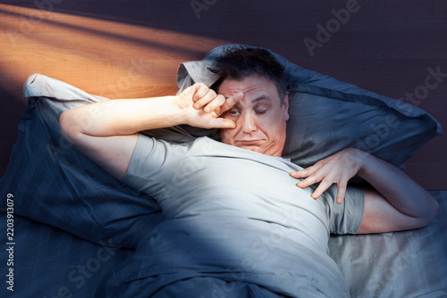 фотографія Tired man waking in bed and opening one eye with his hand as warm morning sunlight enters the bedroom, symbolizing waking up, morning routine, fatigue and the start of a new working day now