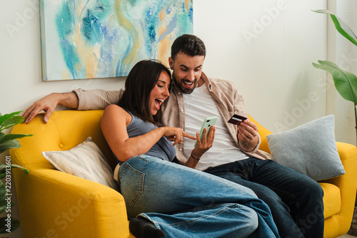A couple lounges on a bright yellow sofa, excitedly scrolling on their phones and sharing a moment of online shopping. The scene captures collaboration, curiosity, and lighthearted joy as they enjoy