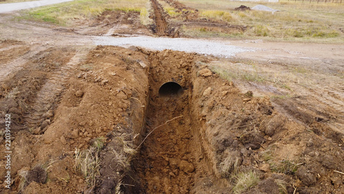 An elevated outdoor photograph capturing a drainage culvert pipe installed inside a newly excavated dirt trench running perpendicular to a dirt road.