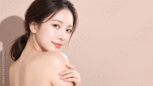 ピンクベージュ背景で肩に手を添えて振り返る日本人女性の透明感ある美容ポートレート