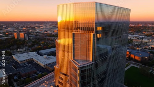 Sunlight glows through glass skyscraper's windows. Aerial view captures golden hour's warm glow. Modern structure dominates urban landscape below. Buildings reflect sun in polished, mirrored surfaces