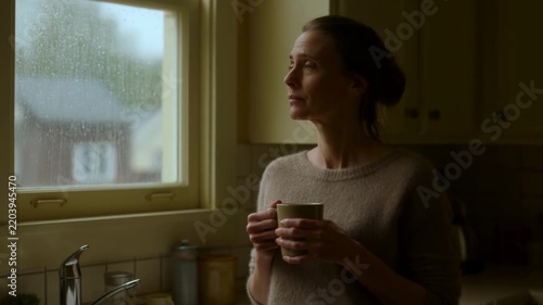 Woman holds mug, gazes out rainy window. Soft light fills cozy kitchen space. Her calm expression suggests quiet reflection. Ideal for scenes of home, rest, or gentle contemplation