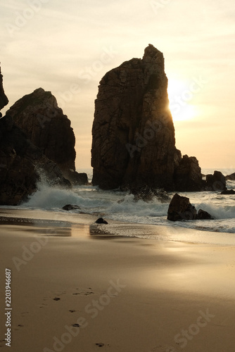 Fotografie Sun shining behind huge sea stack on sandy beach with footprints and crashing wa