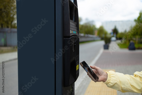 Close up view of a person making a modern contactless payment with a smartphone at an outdoor self-service parking meter. The shot highlights wireless NFC technology, digital banking, urban automation