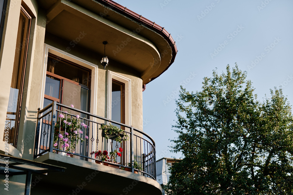 Naklejka premium Cozy house balcony with curved architecture, metal railing and blooming flowers, green tree and blue sky in background. Concept of home comfort and residential lifestyle.