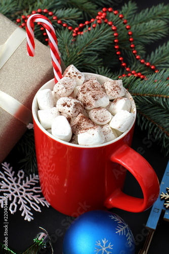 A red mug of cocoa with marshmallows sits on a black background, surrounded by Christmas items.