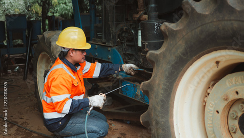 Asian male mechanic applying lubricant spray inspecting tractor machinery in garage.