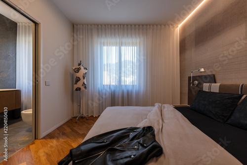 Obraz na płótnie Warm indirect ceiling lighting surrounds an upholstered bed, black leather jacket and sheer curtains beside an open en suite doorway