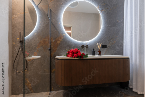 A wide view of an Italian-designed contemporary bathroom pairs a glowing round mirror and curved floating wood vanity with red and pink peonies and roses arranged in the sink.