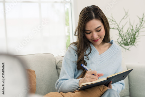 Young Asian woman records daily note in diary book on sofa, relaxed home lifestyle, thoughtful journaling session, weekend planning habit, bright apartment comfort, mindful quiet time indoors.