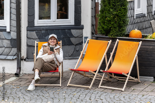 Smiling woman relaxes in deckchair outside slate house in Goslar, enjoying autumn travel mood. Harz tourism, casual fashion and historic streets create cozy lifestyle scene in Germany today
