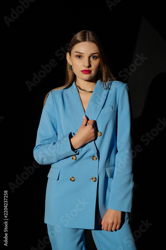Beautiful fashionable woman wearing trendy blue color suit blazer, pants, stylish necklace, posing on black background. Waist up studio fashion portrait