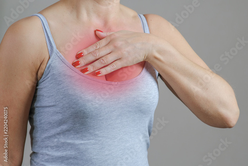 Adult woman holding upper chest with pained facial expression, showing reddened area on skin and discomfort gesture, neutral indoor background, close-up health symptom