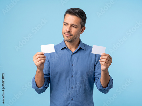 Thoughtful mature man holding two blank white cards making a choice between payment options on blue background
