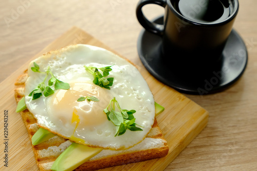 Sunny-side up fried egg on sliced avocado toast with black coffee and parsley on wooden serving board, warm tabletop light, breakfast close view for cafe menus morning