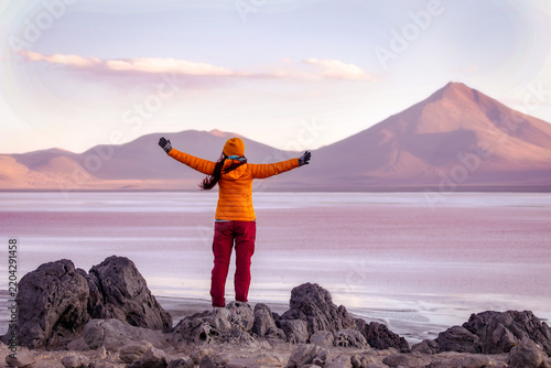 View from behind: a young woman with arms outstretched stands on the shore of Laguna Colorada, Bolivia