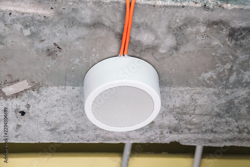 White circular speaker mounted on a raw concrete ceiling, showing exposed orange electrical wiring and industrial design