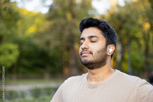 Young man standing outdoors with eyes closed, wearing wireless earbuds and meditating in a park, finding calm, mindful breathing and relaxation amid green, sunlit nature