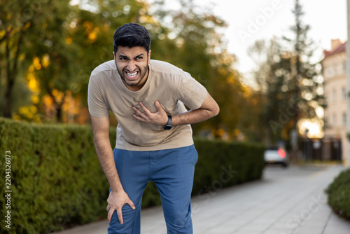Young man experiencing sudden and severe chest pain, grabbing his chest and bending over in agony while standing on a sidewalk outdoors, indicating a potential heart attack or medical emergency
