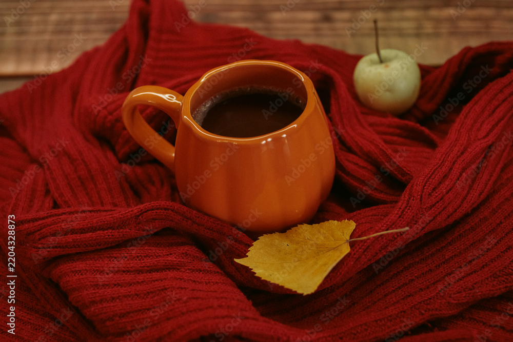 Fototapeta premium An orange mug of coffee resting on a red knitted sweater beside an open book and a yellow autumn leaf, with soft film grain, high quality photo