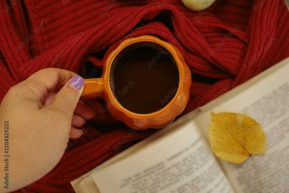 Fototapeta premium An orange mug of coffee resting on a red knitted sweater beside an open book and a yellow autumn leaf, with soft film grain, high quality photo