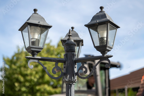 Obraz na plátně Close-up of traditional black outdoor lamps mounted on an ornate metal post, with trees and a blue sky in the background