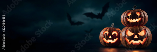 A spooky halloween composition with three carved, glowing jack-o-lanterns stacked and placed on a dark surface against a cloudy night sky with bats
