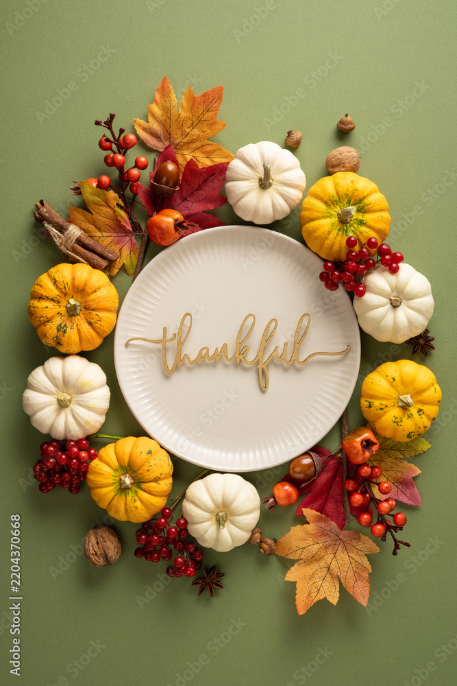 Fototapeta premium Thankful plate surrounded by pumpkins, autumn leaves and seasonal spices for Thanksgiving Day celebration on vertical green tabletop