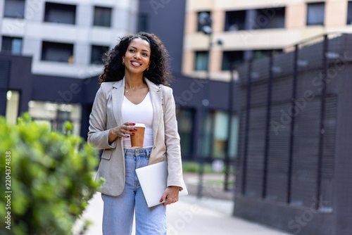 Smiling professional woman walking outdoors with takeaway coffee and laptop, representing commuting, career mobility and a modern urban work lifestyle