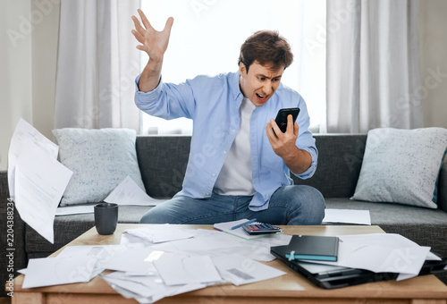 Angry man throwing paper bills and shouting at smartphone screen. Financial stress, high utility costs, inflation, unpaid overdue bills, rising energy prices, economic crisis.