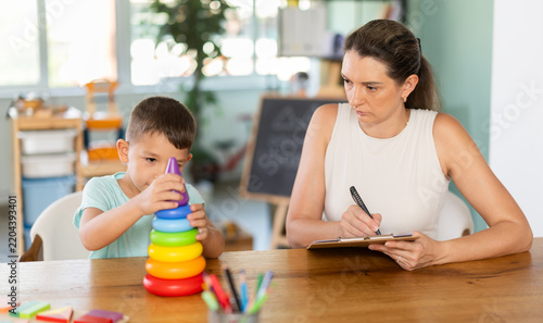 Professional child psychologist observing little boy assembling toy pyramid on table at development center