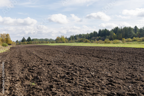 Ploughed dark soil field ready for planting crops with green meadows and pine forest under a cloudy blue sky.