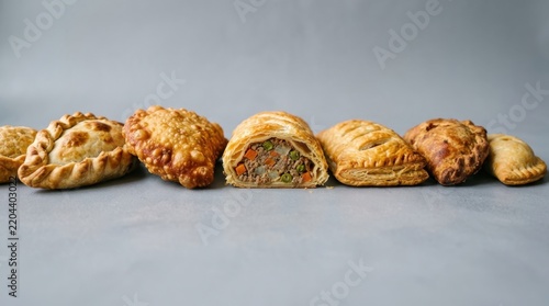 Variety of golden brown savory pastries and empanadas on a gray surface, including a sliced puff pastry roll filled with minced meat and vegetables, perfect for culinary and bakery themes.