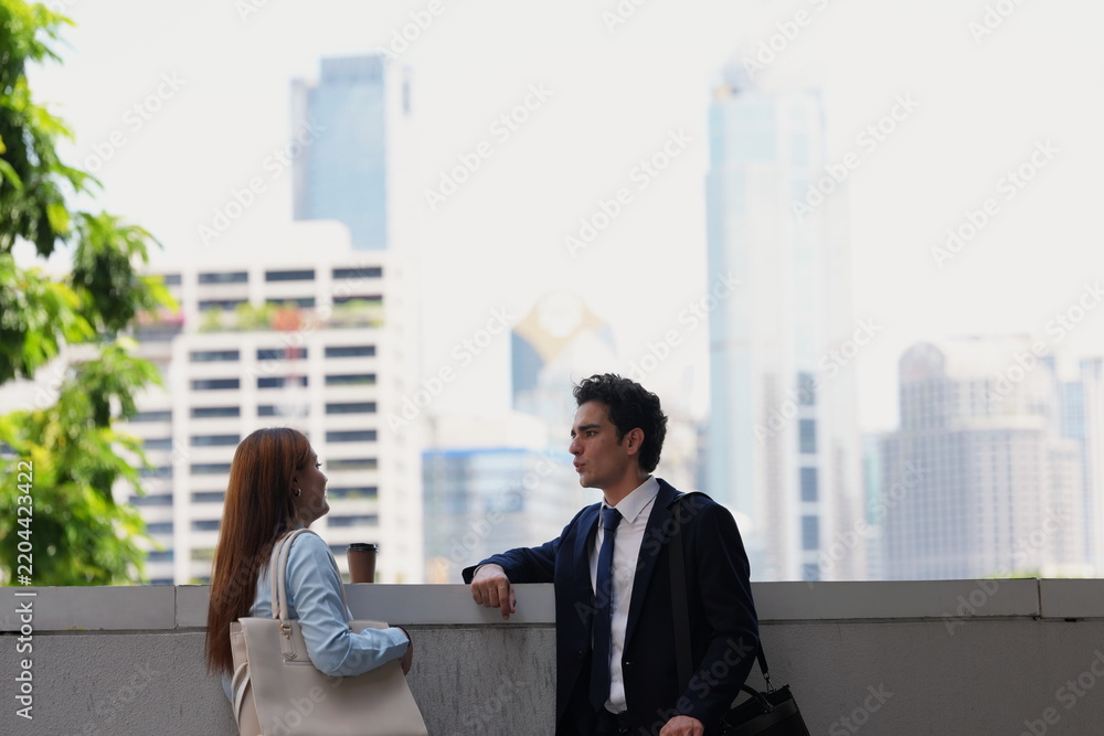 Obraz premium Two young professionals take a coffee break at a parapet overlooking the city, a strong skyline image for real estate, careers and lifestyle campaigns, with hazy towers behind
