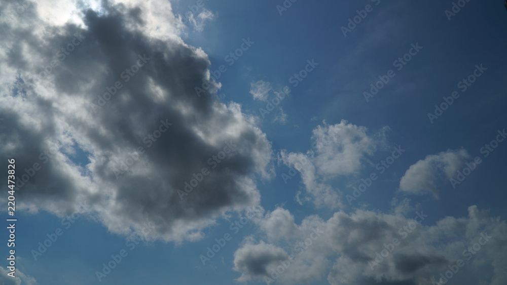 Obraz premium Dramatic Sun Peeking Above Dark Cloud in Blue Sky with Scattered Clouds