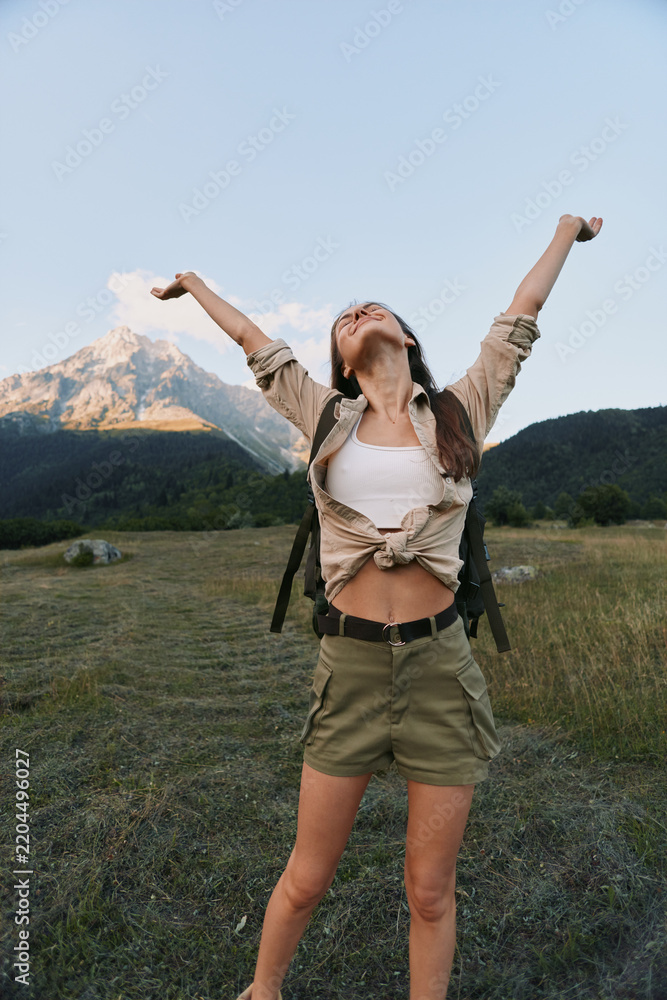 Obraz premium Young woman embraces outdoor freedom, raising arms in triumph against mountains, standing in an open field with a clear sky, expressing carefree lifestyle and travel vibe