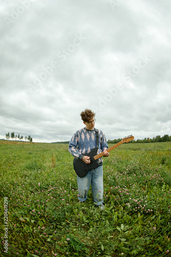 Vertical image of Caucasian young adult musician playing electric guitar in rural field. Visual for indie music production, creative youth lifestyle, and media advertising