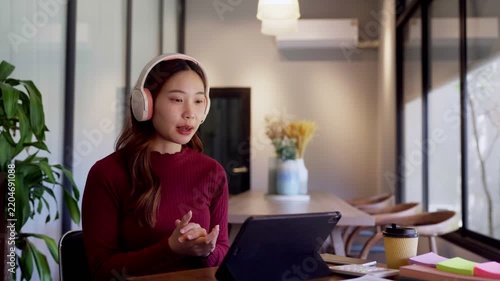 Young Asian woman wearing headphones gestures during video call on tablet in modern office