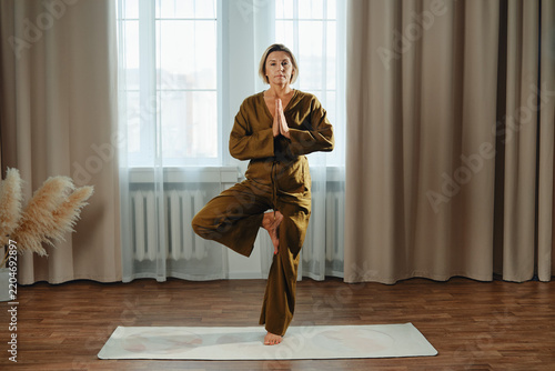 Middle-aged woman stands on one leg with hands in prayer position in indoor space. Natural light enters through large windows. Simple room has wooden floor and soft curtains.